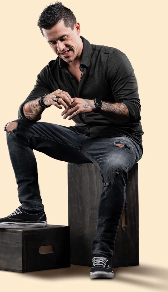 Johnny Destro sentado numa caixa de madeira, camisa preta, sorrindo